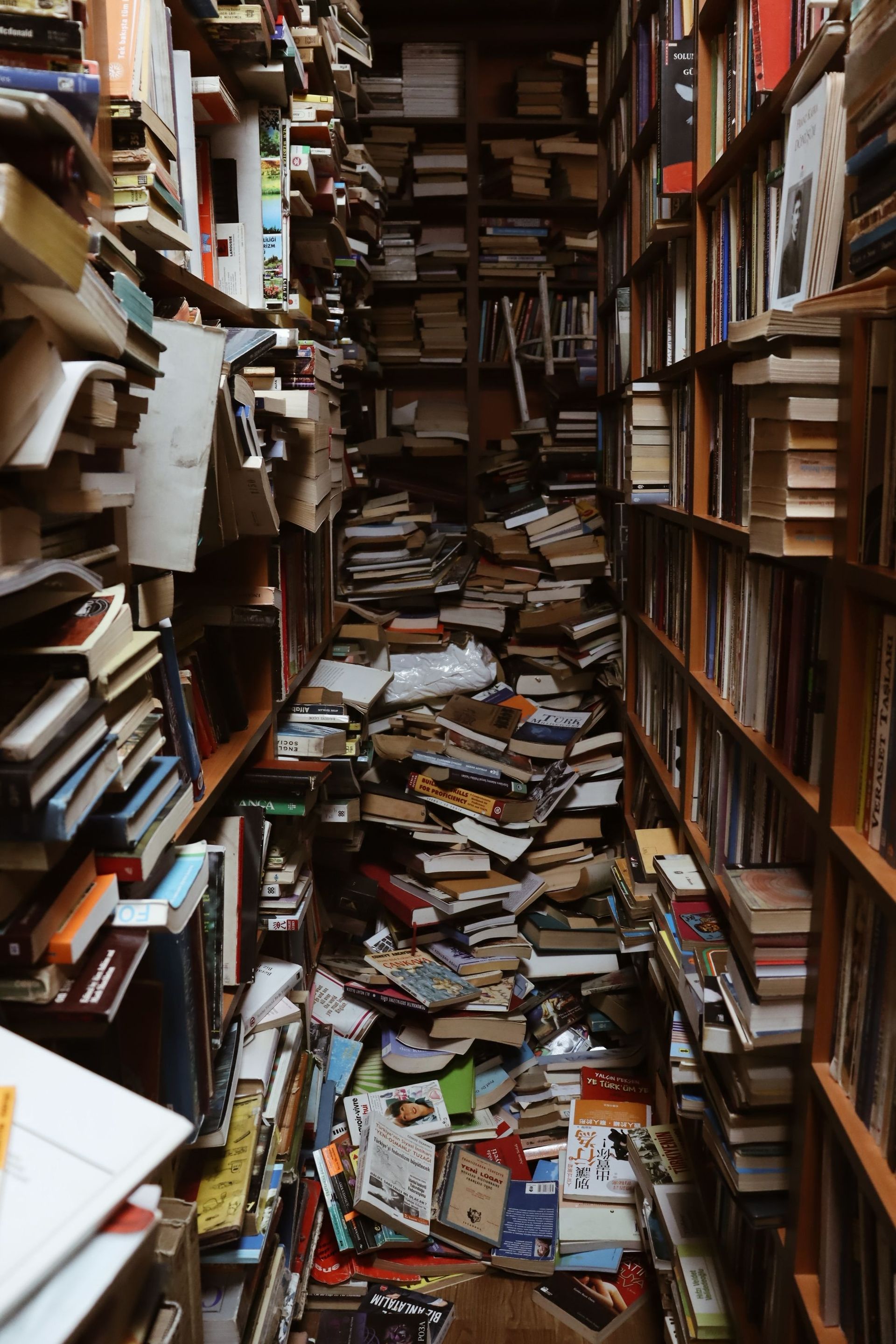 Overflowing bookshelves in a narrow library aisle creating a chaotic atmosphere with scattered books.